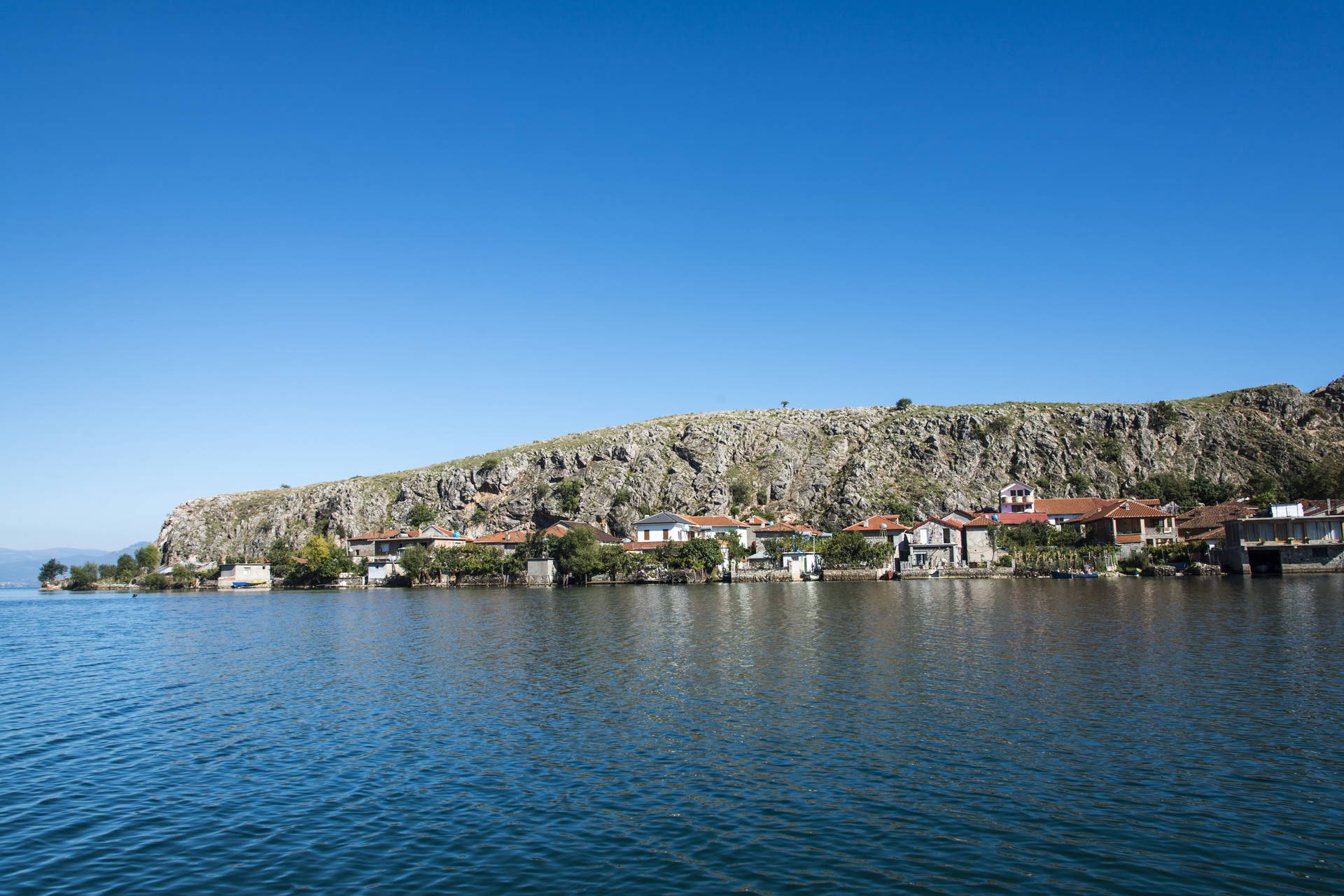 Linia brzegowa z domami, skaliste wzgórze i spokojna woda pod błękitnym niebem.