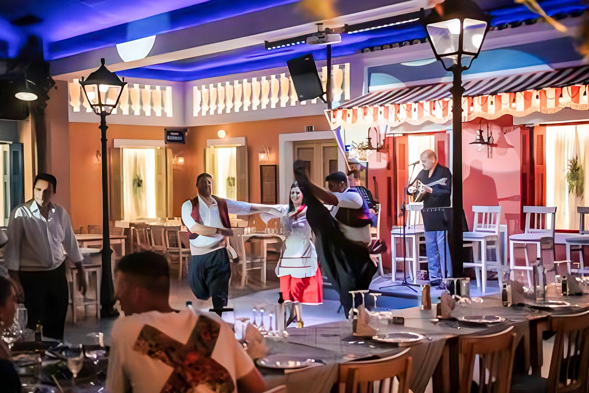 Tancerze w strojach ludowych występują w greckiej restauracji. W tle widać muzyka i stoły przygotowane dla gości.