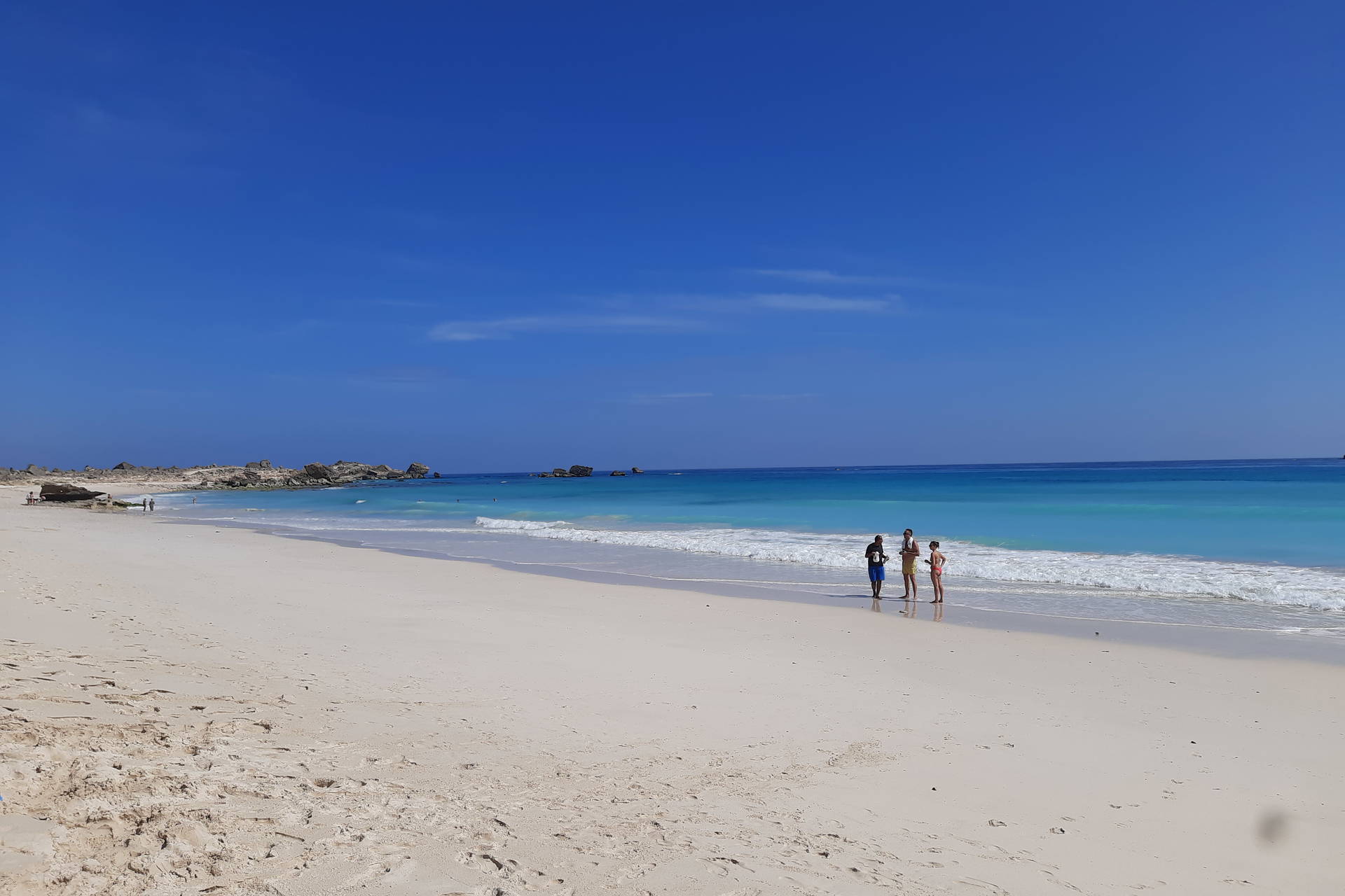 Trzy osoby stoją na piaszczystej plaży. W tle widać błękitne morze i niebo.
