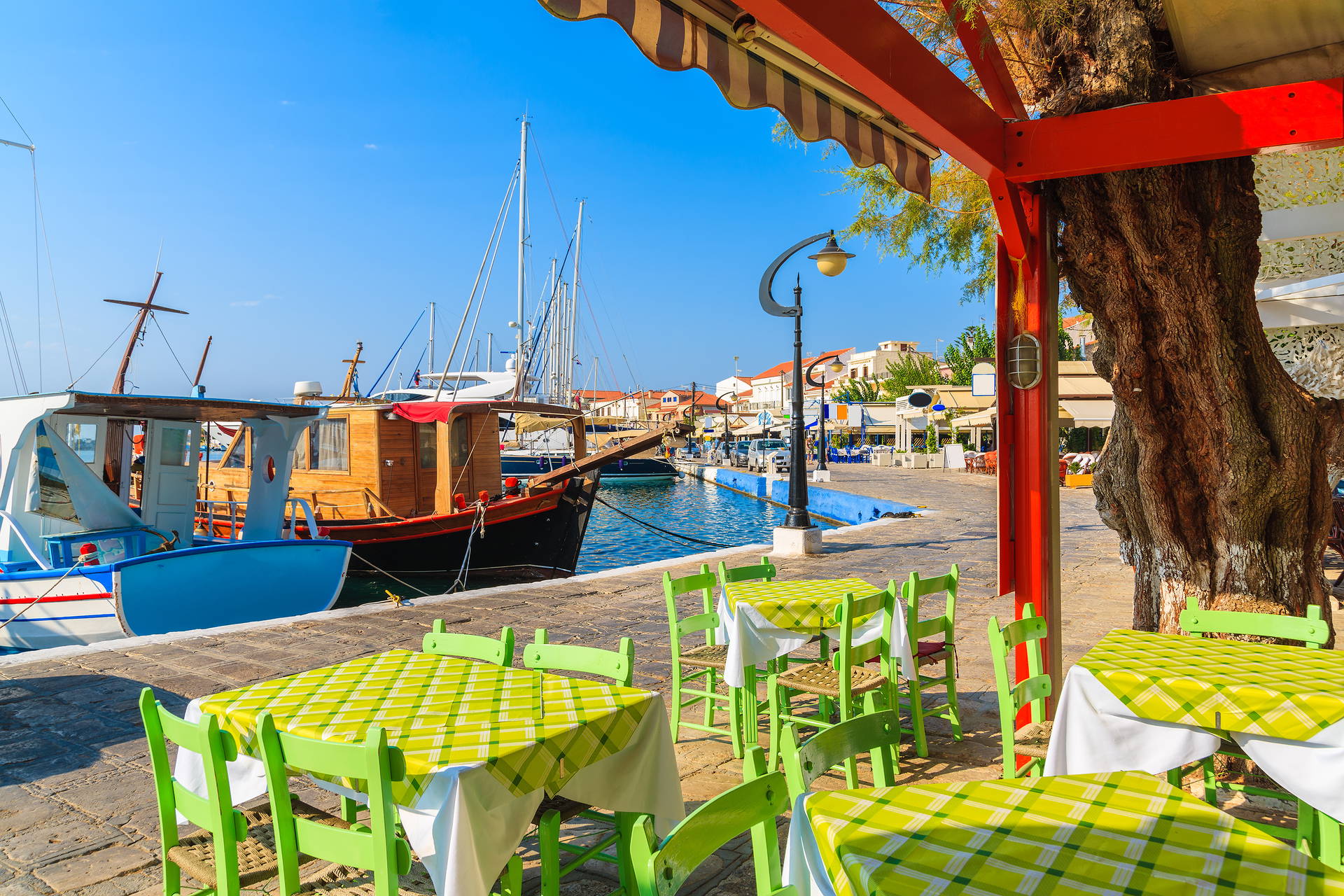 Restauracja z widokiem na port. Na zewnątrz zielone stoliki. W tle zacumowane łodzie.