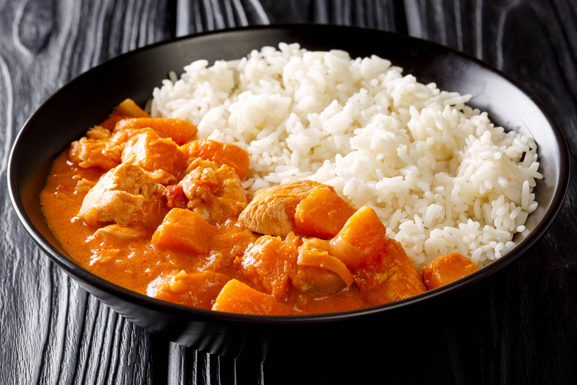 Czarna miska z curry z kurczakiem i warzywami podana z porcją białego ryżu na ciemnym, drewnianym stole.