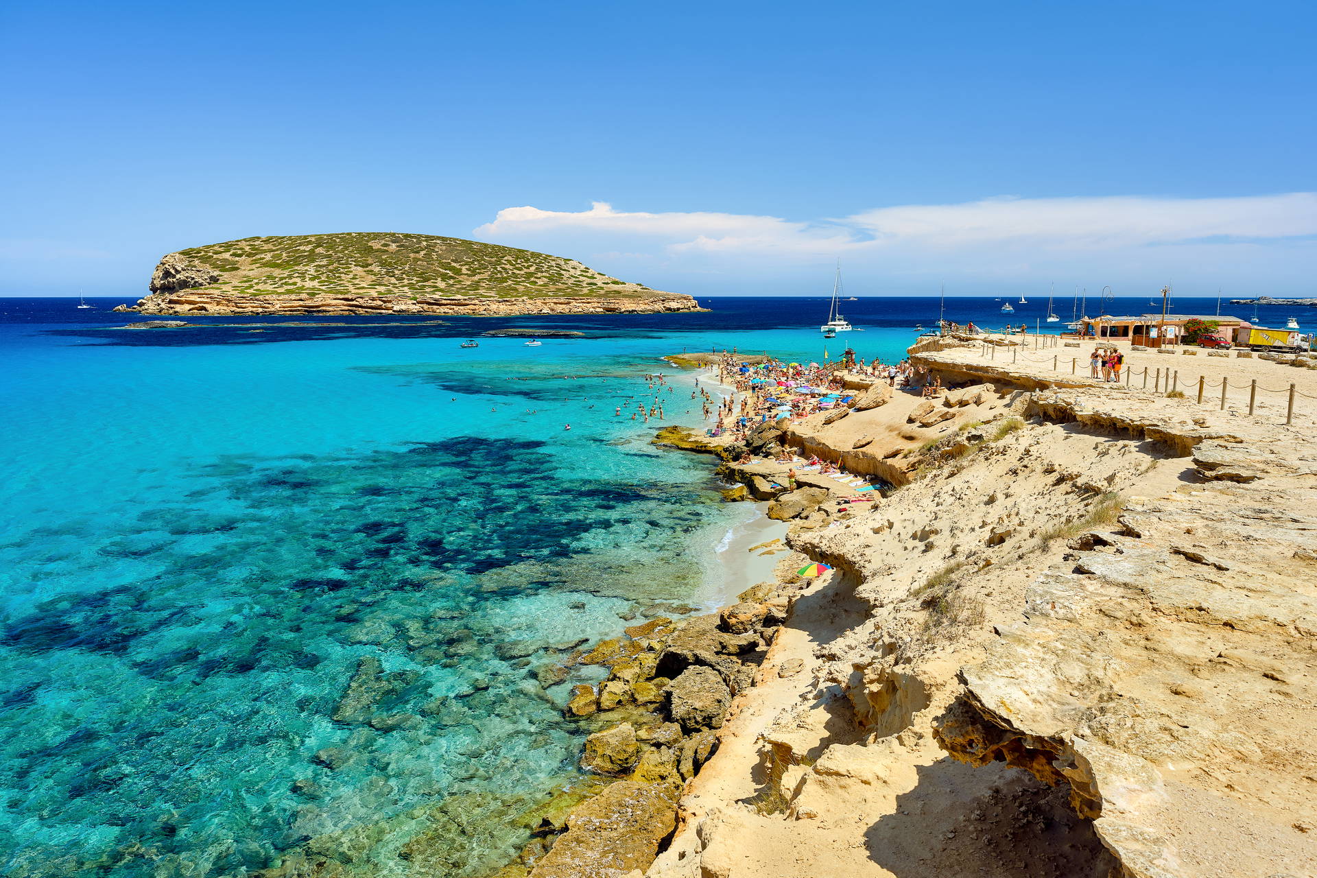 Plaża Cala Comte na Ibizie: skaliste wybrzeże, tłum ludzi, turkusowe morze i mała wyspa w oddali.