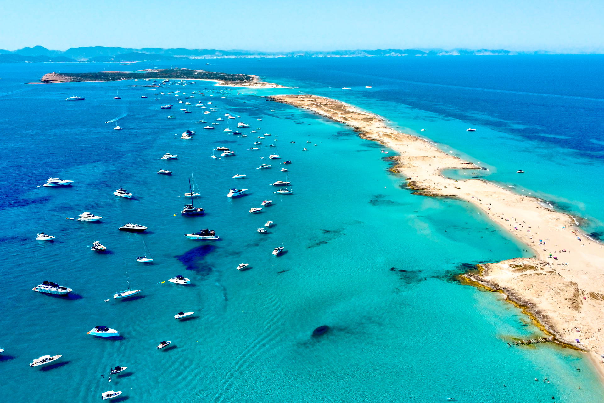 Plaża Ses Illetes na Formenterze z lotu ptaka. Wokół piaszczystego cypla, na turkusowej wodzie, cumuje wiele jachtów.