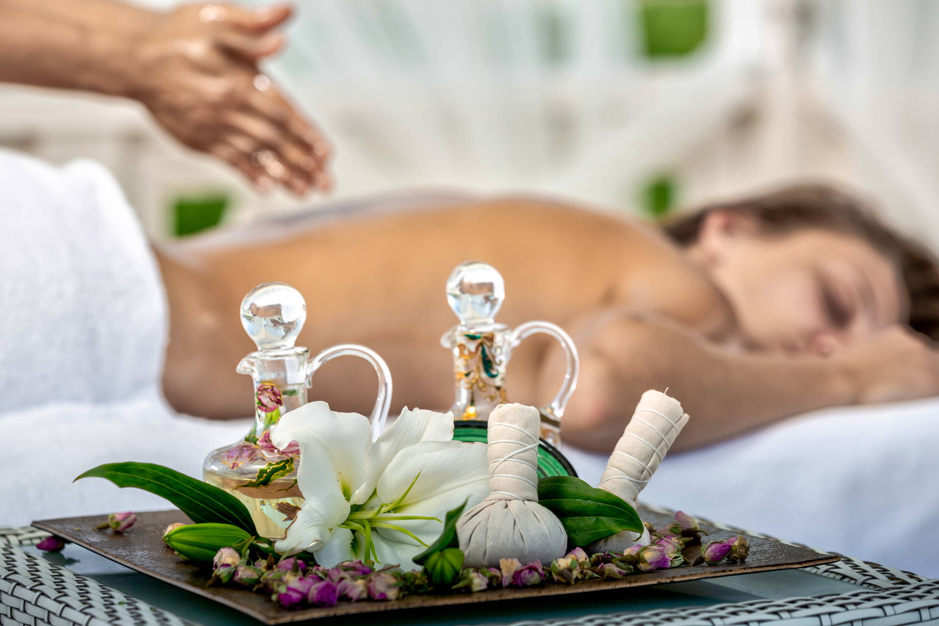 Relaksujący masaż pleców. Na pierwszym planie taca z olejkami, stemplami ziołowymi i kwiatem lilii.