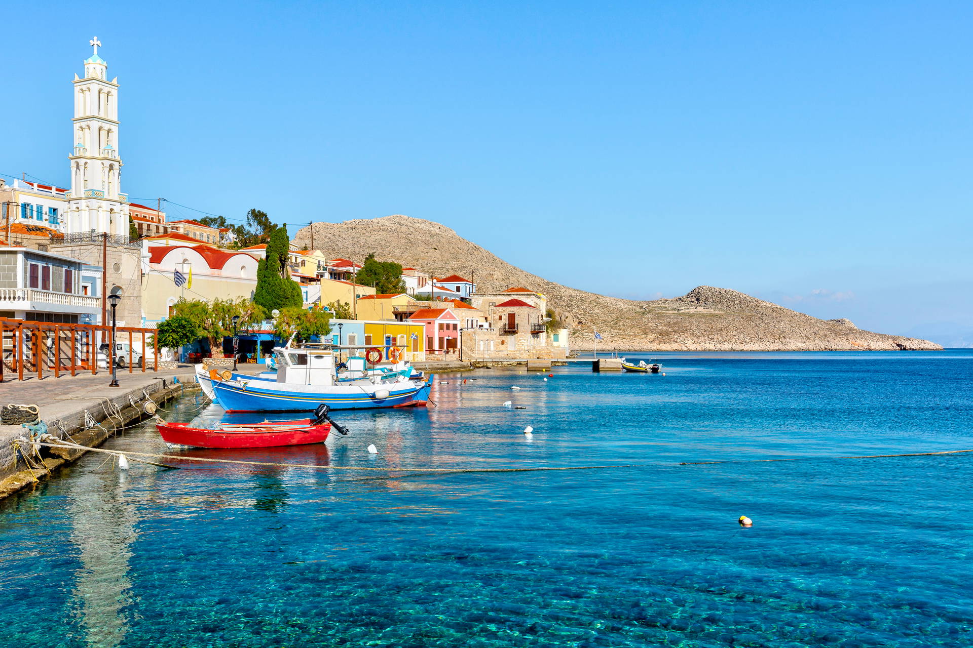 Mały port na greckiej wyspie Halki. Na turkusowej wodzie kołyszą się łodzie, a w tle widać kolorowe budynki miasteczka i białą dzwonnicę.
