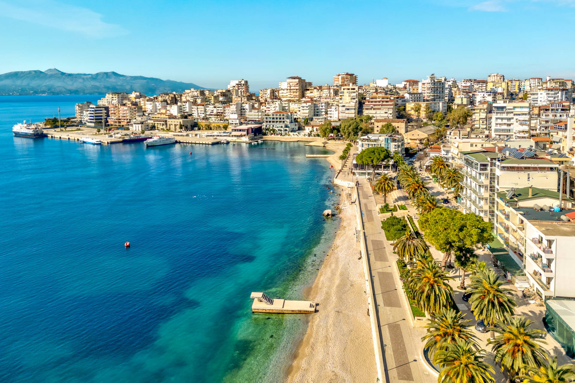 Saranda w Albanii z lotu ptaka. Wzdłuż wybrzeża widać lazurowe morze, plażę, promenadę z palmami i gęstą miejską zabudowę.