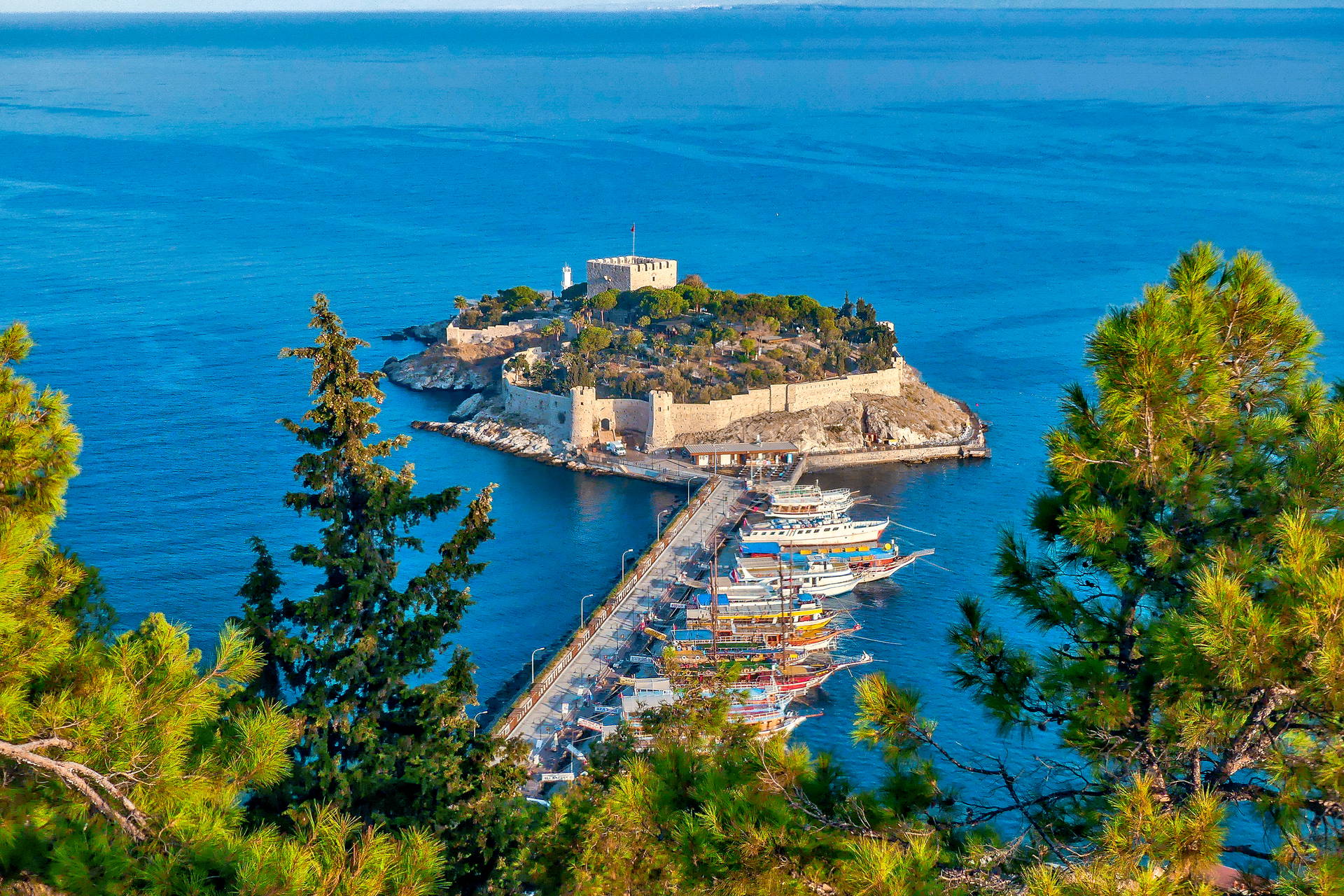 Widok z góry na Zamek Güvercinada w Kuşadası, molo i łodzie na tle błękitnego morza.