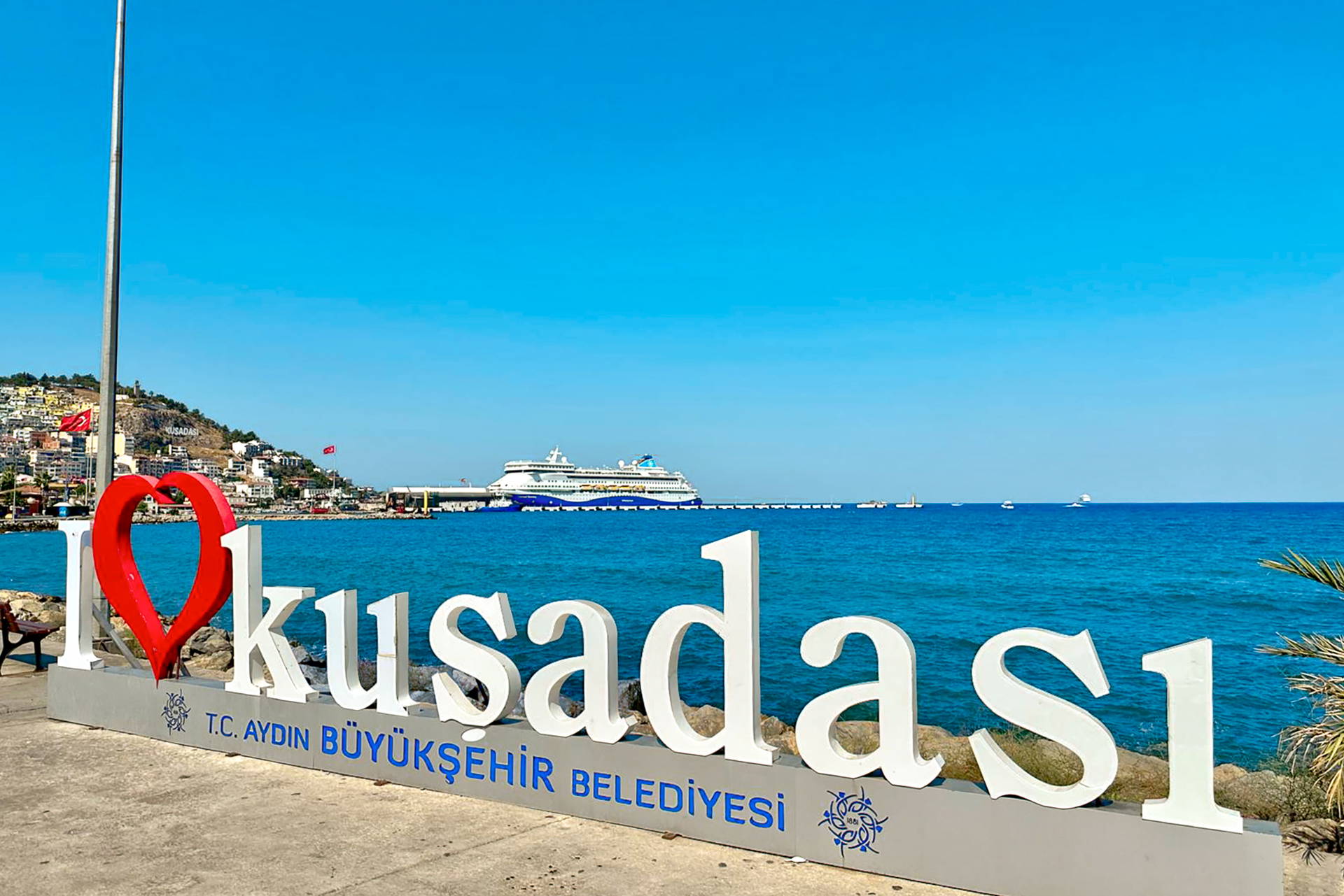 Napis "I love Kuşadası" na nadbrzeżu. W tle błękitne morze i statek wycieczkowy w porcie.