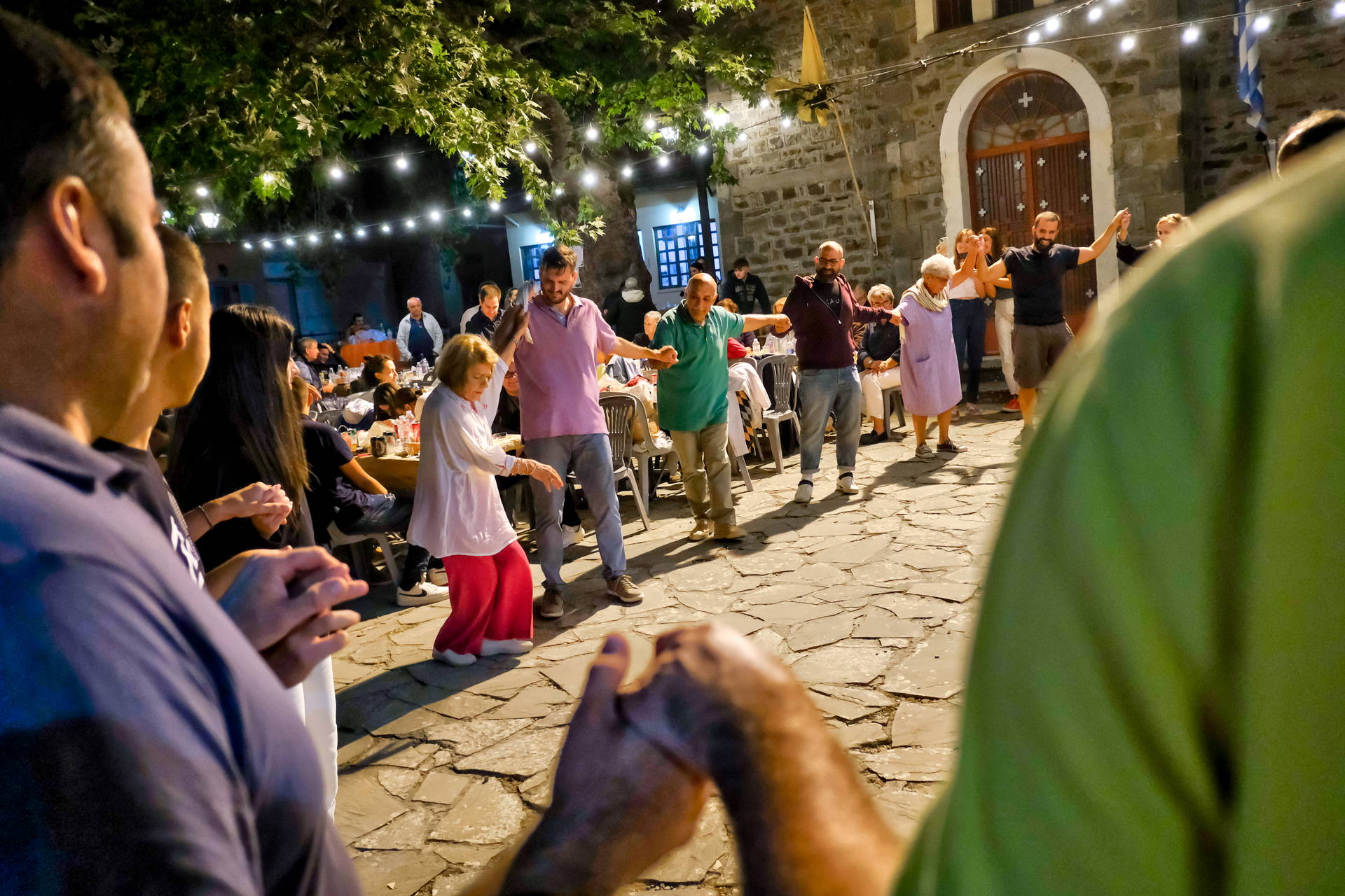 Grupa ludzi tańczy w kręgu tradycyjny grecki taniec na kamiennym placu, oświetlonym nocą.