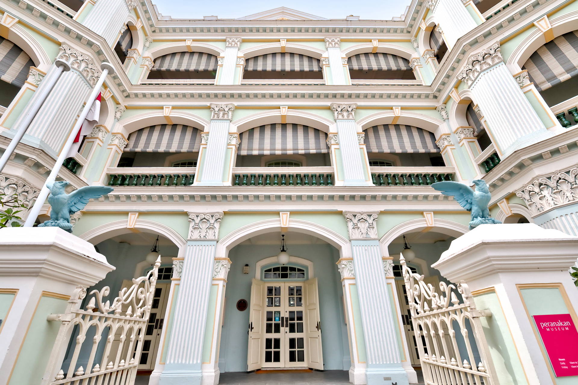 Pastelowa fasada Muzeum Peranakan z kolumnami, łukami oraz ozdobną bramą, której pilnują rzeźby orłów.