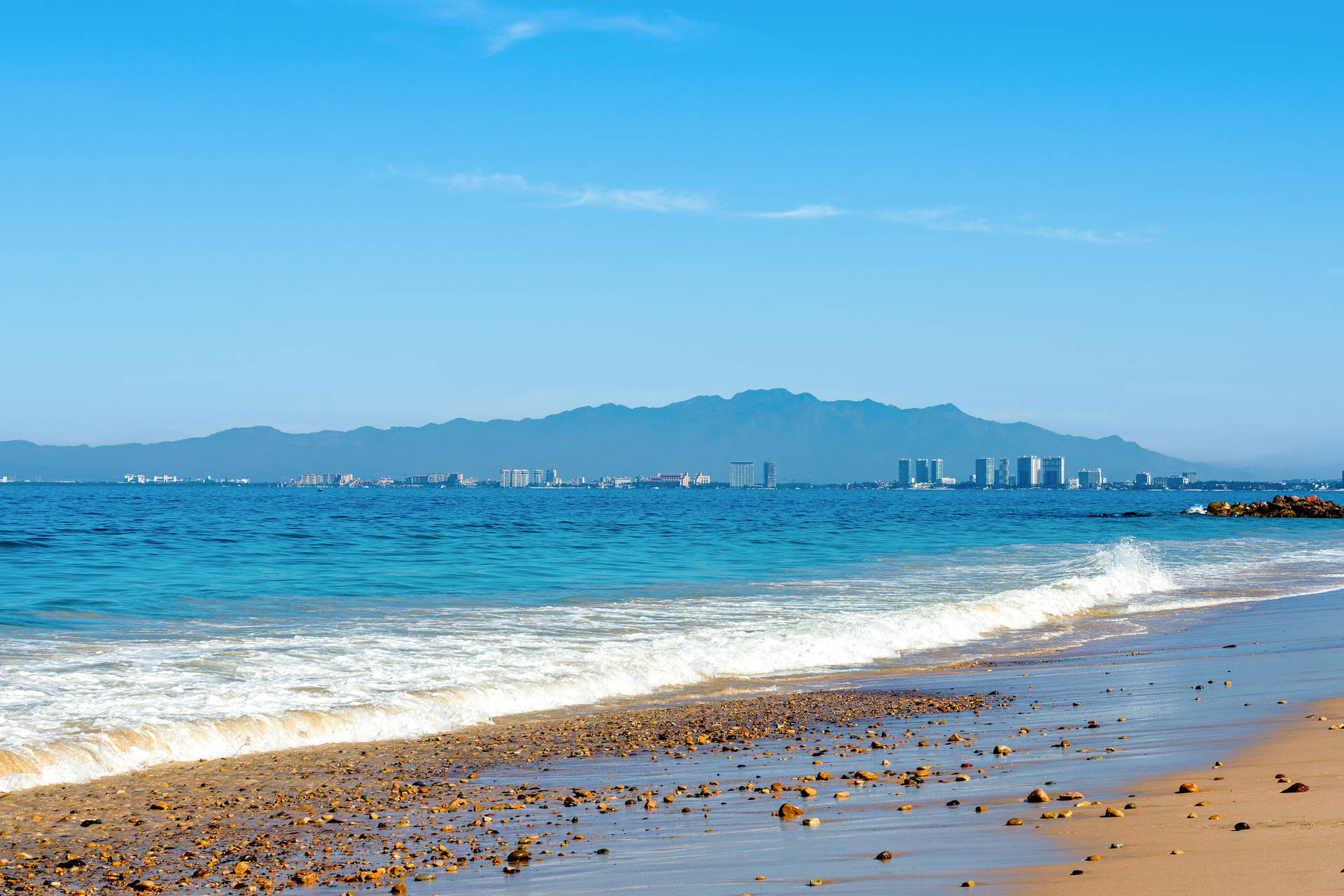 Lazurowe fale oceanu na plaży w Puerto Vallarta, z panoramą miasta i pasmem górskim w tle.