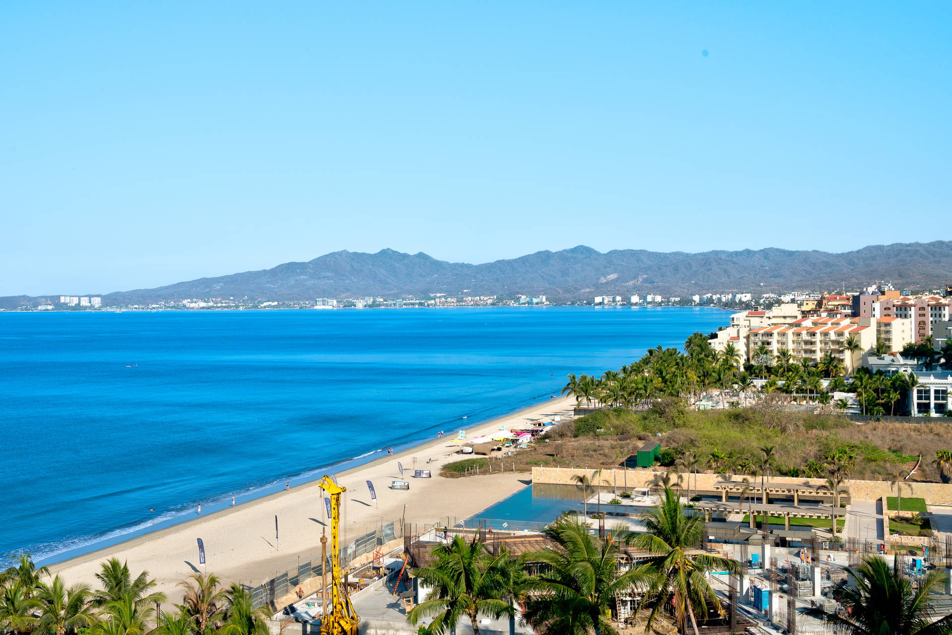 Panoramiczny widok na wybrzeże Puerto Vallarta. Na pierwszym planie szeroka plaża i błękitny ocean, w tle hotele oraz góry.