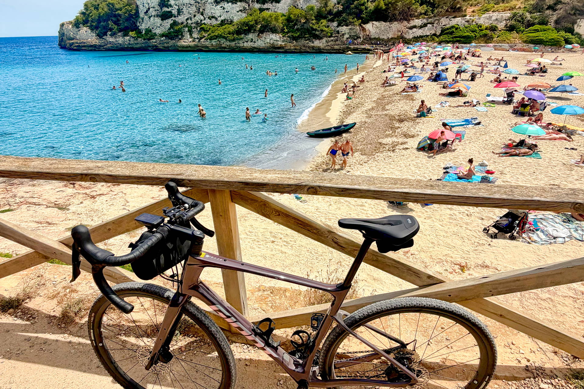 Rower gravelowy na tle zatłoczonej plaży Cala Llombards z turkusową wodą.
