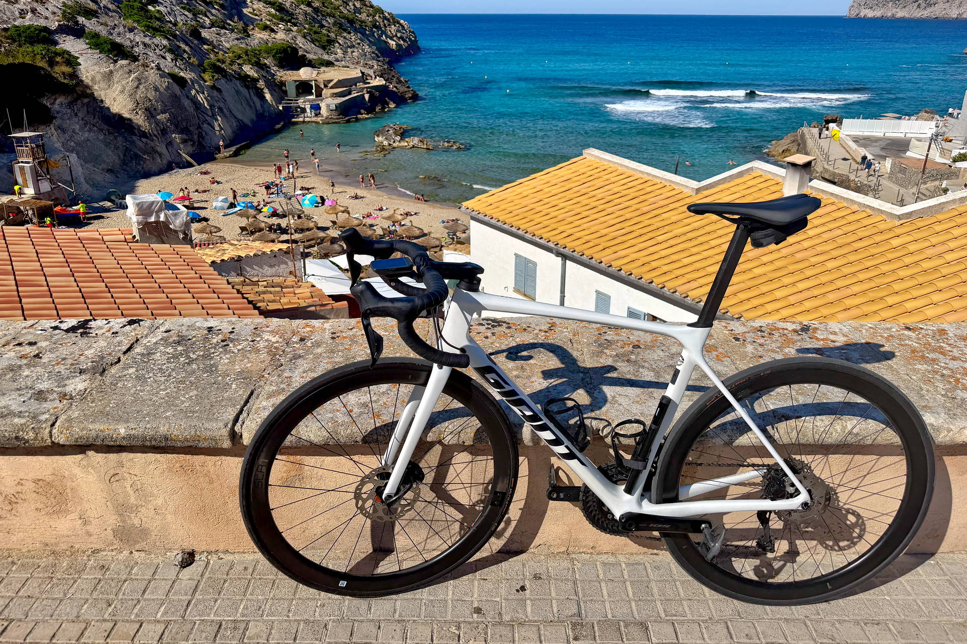 Biały rower szosowy na murku z widokiem na plażę i zatokę Cala Deià na Majorce.