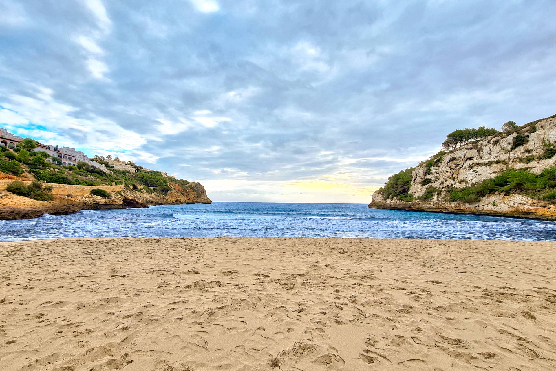 Piaszczysta plaża w zatoce Cala Romantica na Majorce, otoczona skalistymi klifami porośniętymi zielenią.