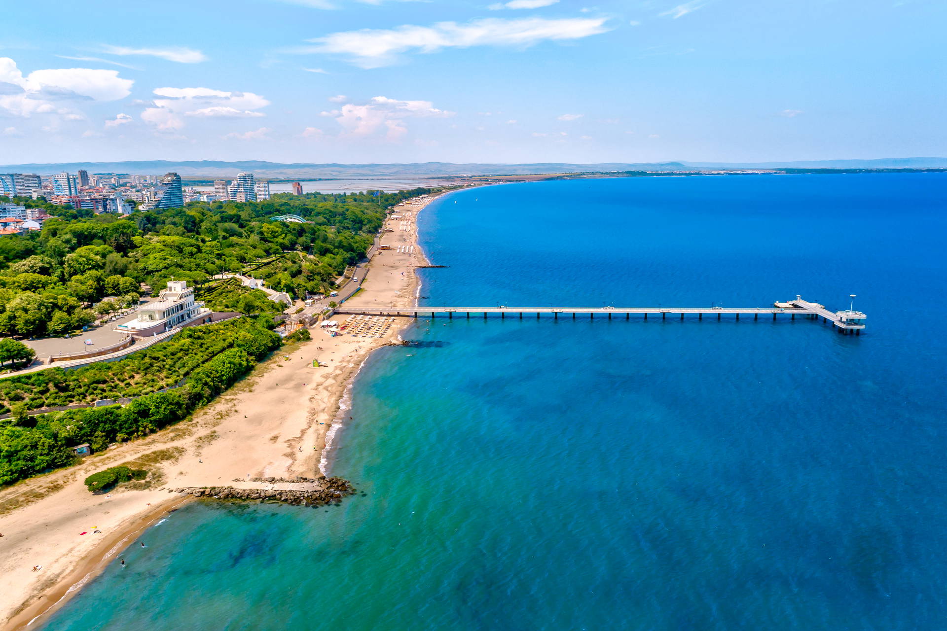 Widok z lotu ptaka na molo w Burgas, które wychodzi z piaszczystej plaży w głąb błękitnego morza. W tle panorama miasta.