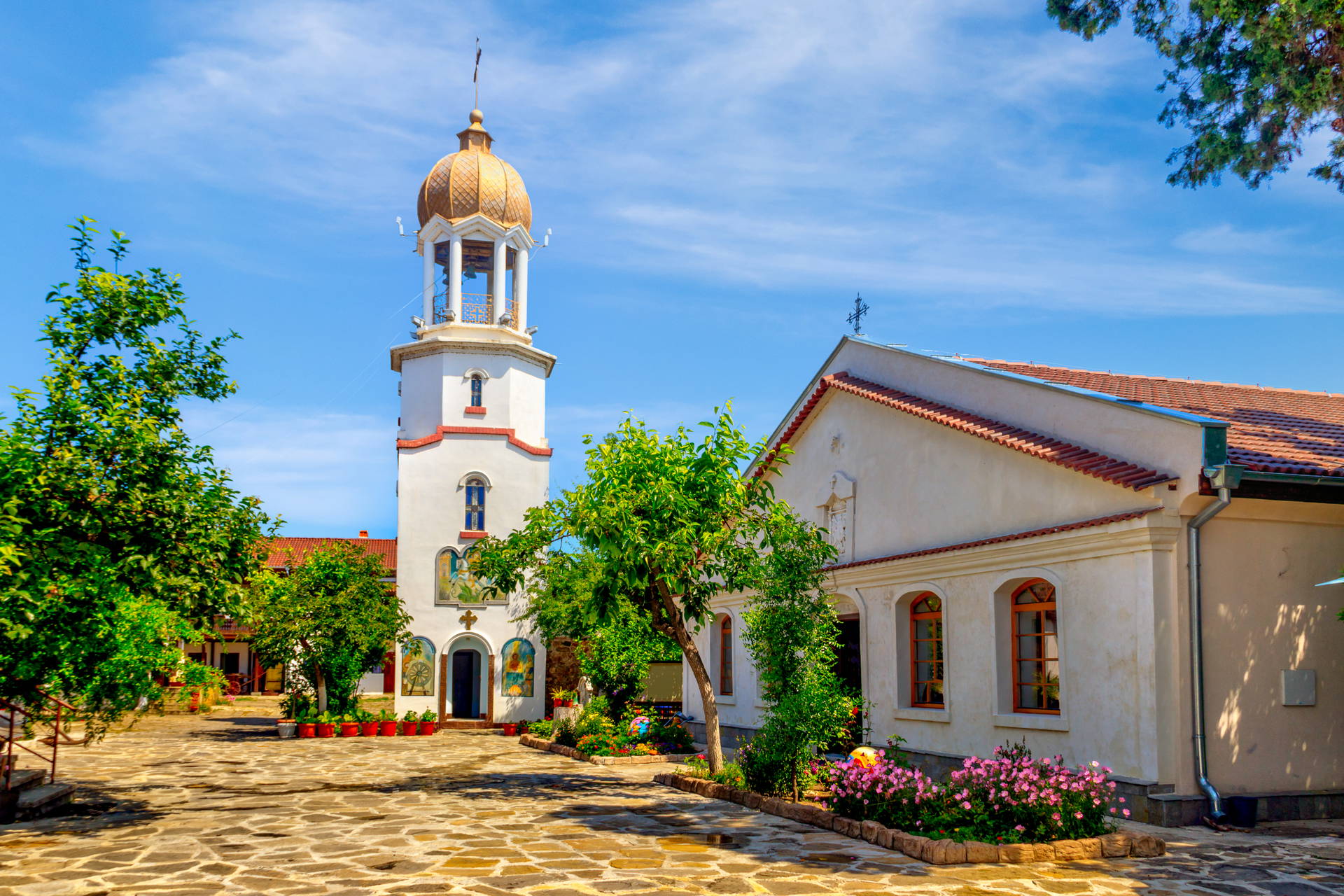 Klasztor św. Jerzego w Pomorie. Biała dzwonnica ze złotą kopułą i budynek kościoła na kamiennym dziedzińcu, wśród zieleni.