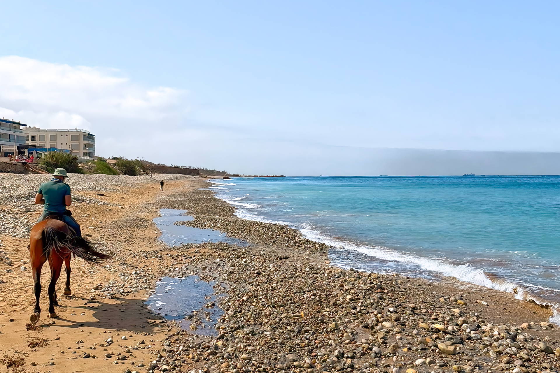 Jeździec jedzie konno wzdłuż kamienistej plaży nad turkusowym morzem w słoneczny dzień.