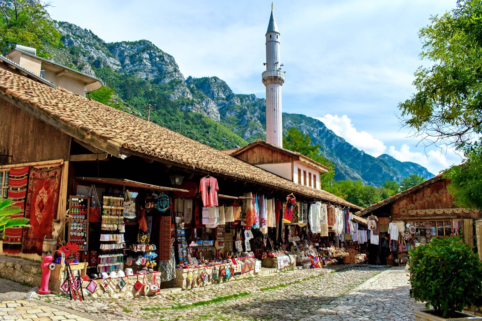 Brukowana uliczka Starego Bazaru w Kruji w Albanii. Po bokach stoją stragany z pamiątkami, a w tle widać minaret i góry.
