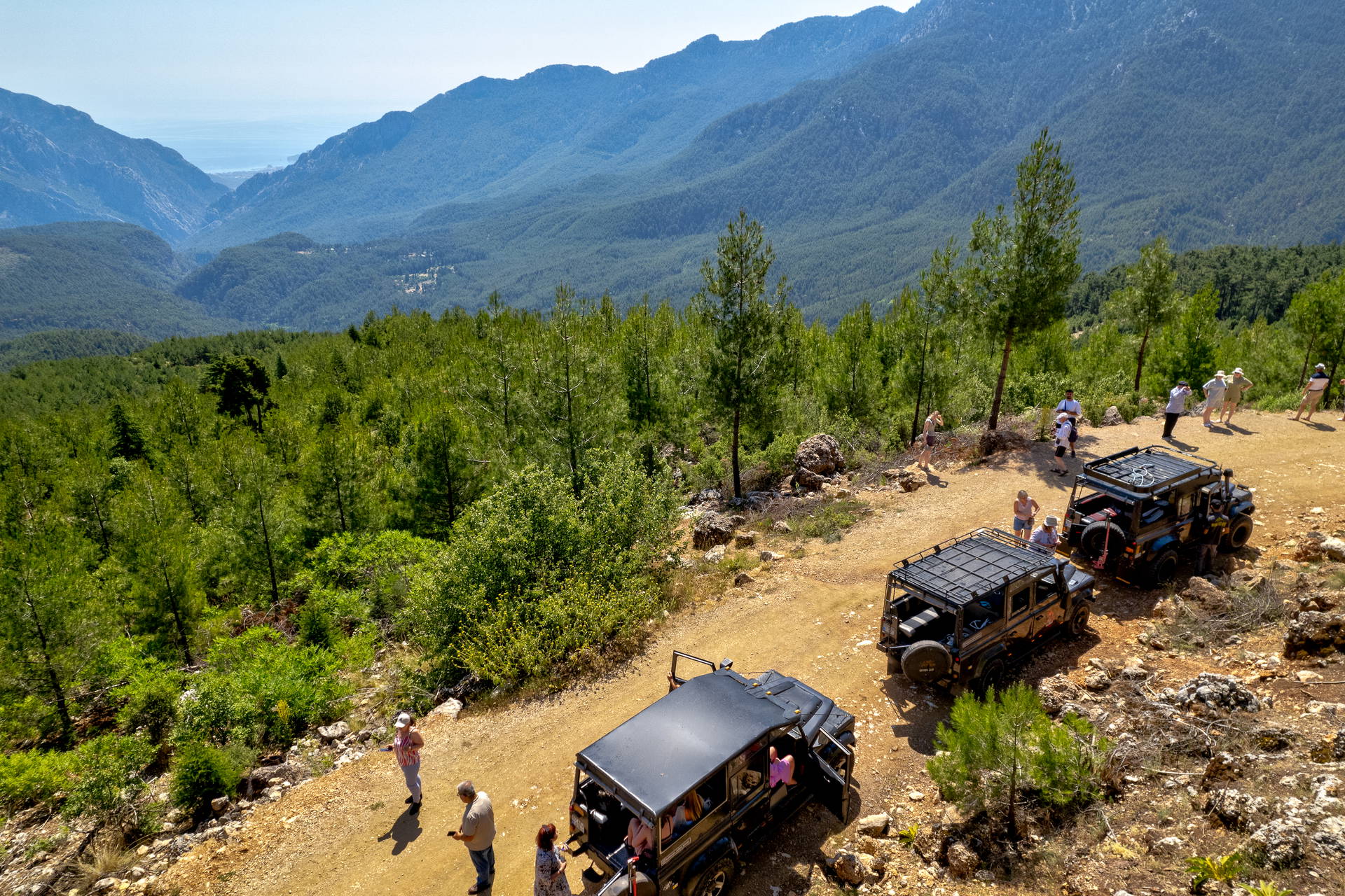 Turyści i dwa samochody terenowe na postoju podczas jeep safari w górach, z widokiem na zalesioną dolinę.