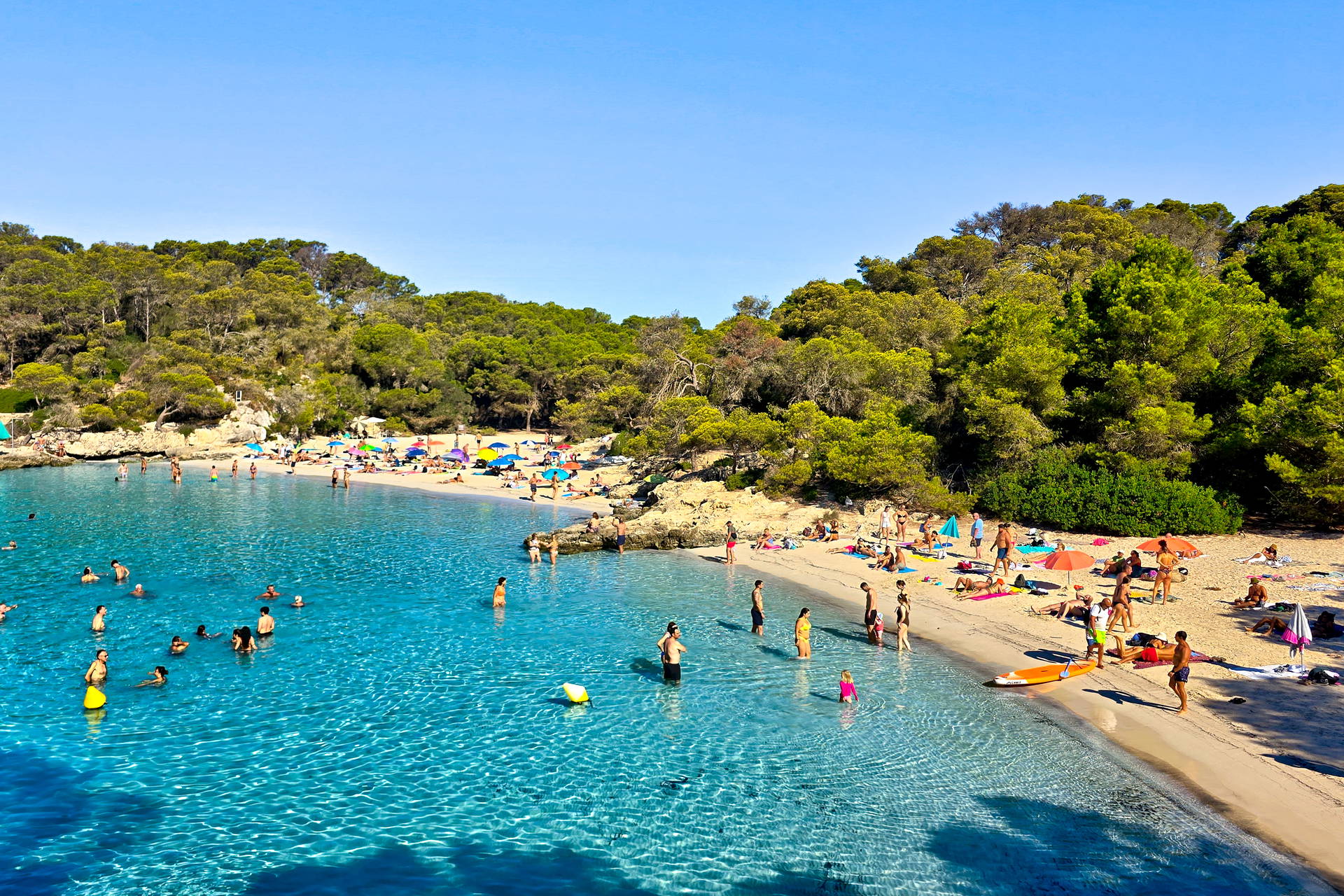 Słoneczna plaża Cala Agulla na Majorce, pełna ludzi, którzy opalają się i kąpią w turkusowej wodzie.