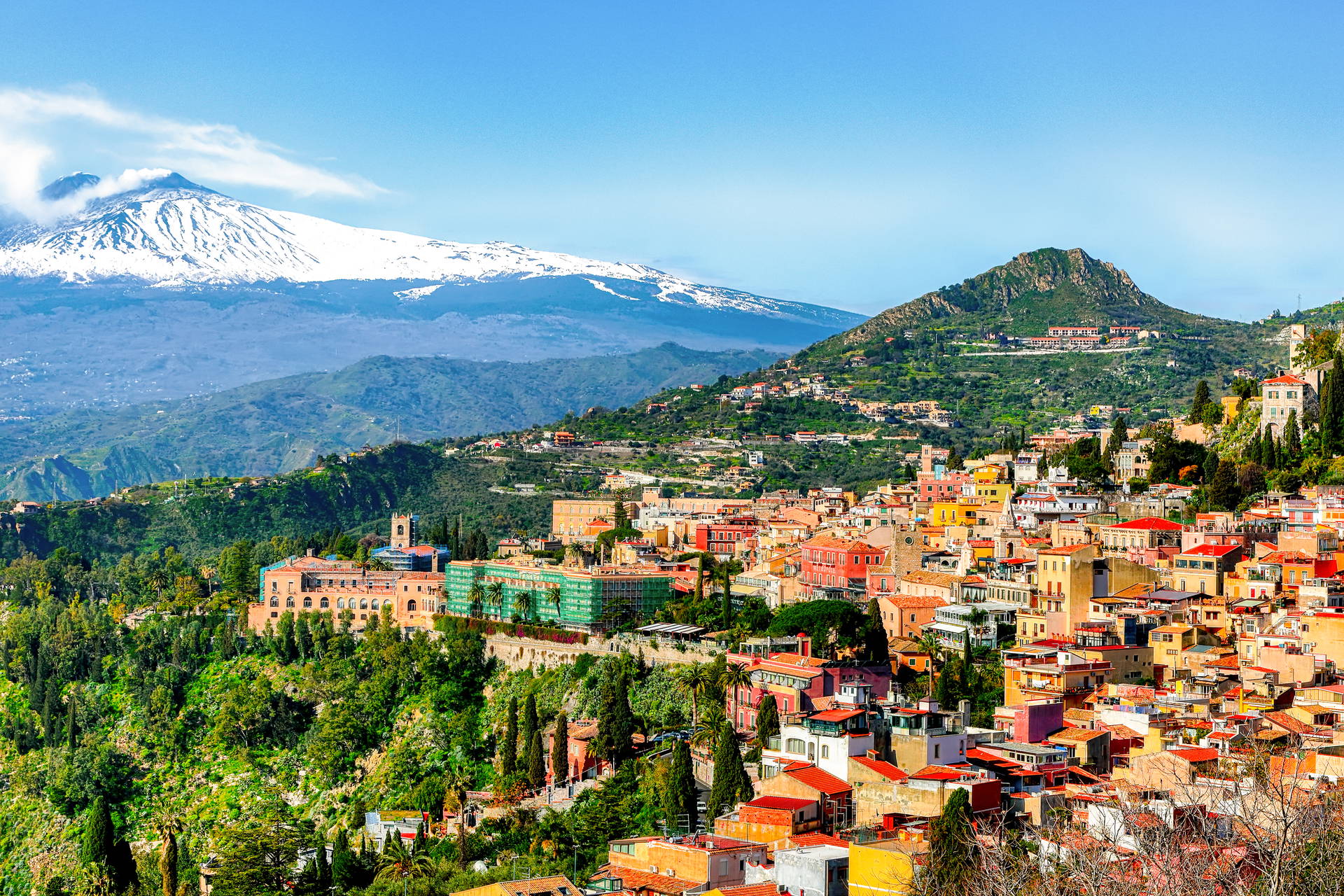 Widok na miasto Taormina na Sycylii: budynki na zboczu góry oraz ośnieżony wulkan Etna w tle.