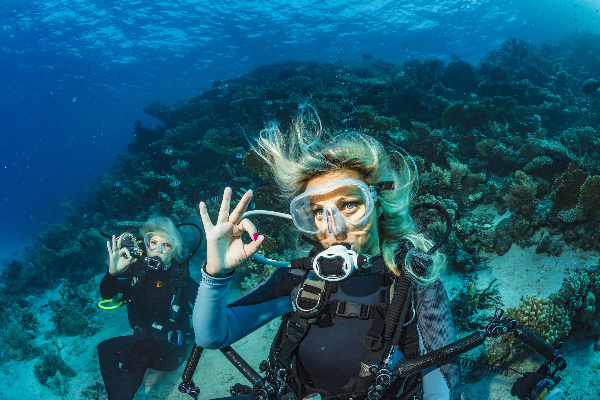 Dwie kobiety nurkują w błękitnym oceanie i pokazują znak "OK".