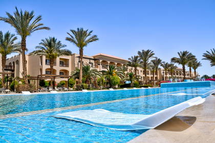 Basen hotelowy z leżakami, palmami i budynkami w tle.