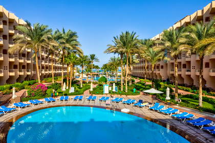 Kompleks wypoczynkowy z basenem, palmami i budynkami hotelowymi w tle.