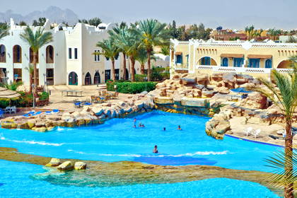 Kompleks hotelowy z basenem, palmami i górami w tle.