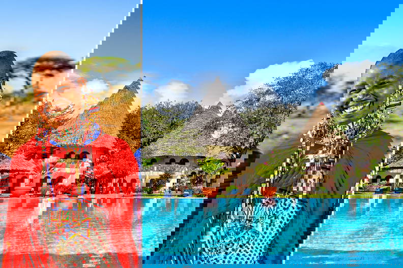 Kobieta z plemienia Masajów po lewej stronie kadru. Po prawej basen i budynki w afrykańskim stylu.