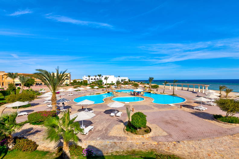 Hotel z basenami, leżakami i palmami. W tle widok na morze. Zdjęcie z góry.