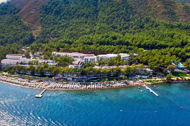 Widok z lotu ptaka na plażę, budynki kurortu i zalesione wzgórza w tle.