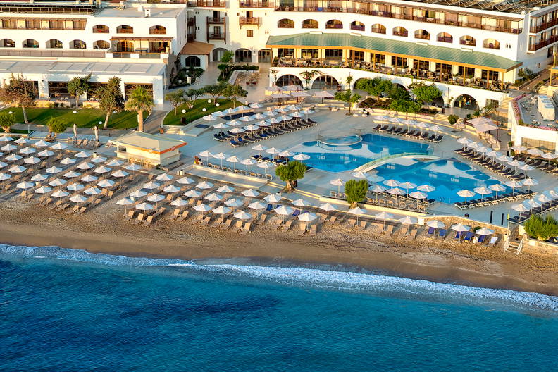 Widok z góry na duży hotel z basenem przy piaszczystej plaży z leżakami i błękitnym morzem.