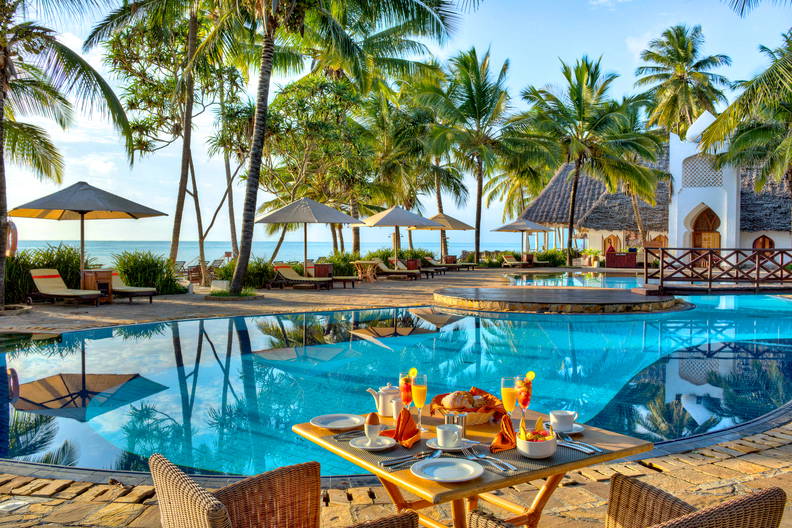 Stół śniadaniowy na tle basenu z palmami i leżakami.