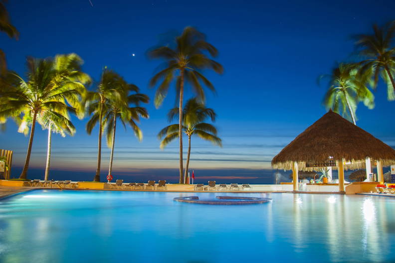 Oświetlony nocą basen z palmami i barem pod strzechą.