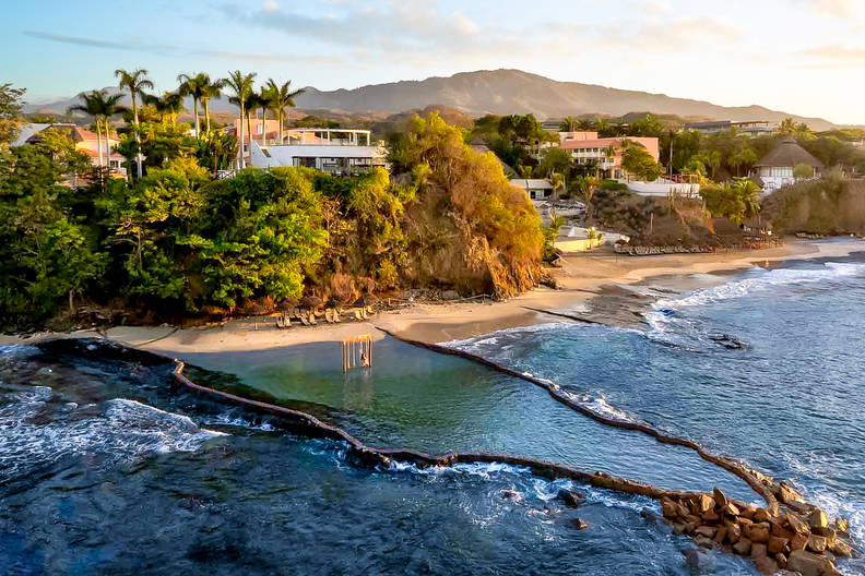 Widok z lotu ptaka na plażę, zabudowania i skalną barierę w oceanie.