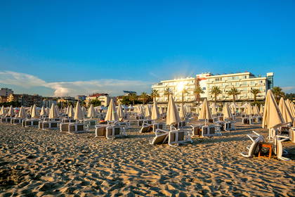 Plaża z leżakami, parasolami i budynkiem hotelu w tle.