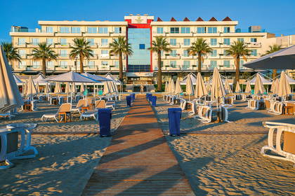 Hotel na plaży z palmami, leżakami i drewnianą ścieżką.