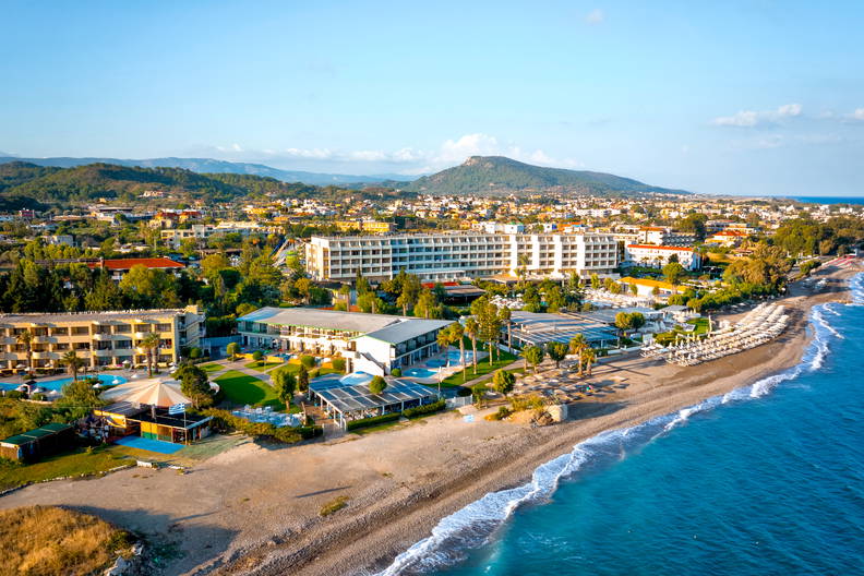 Widok z lotu ptaka na kompleks hotelowy przy kamienistej plaży, z miastem i górami w tle.