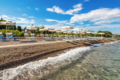 Słoneczna, kamienista plaża z rzędami leżaków i parasoli. Fale rozbijają się o brzeg.