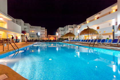 Nocny widok na hotelowy basen, leżaki i budynki w tle.