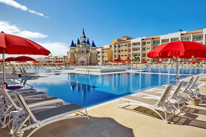 Duży basen hotelowy z leżakami i czerwonymi parasolami na tle budynku stylizowanego na zamek.