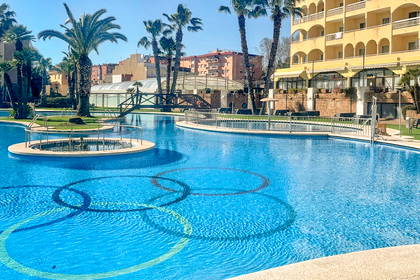 Duży, słoneczny basen hotelowy z kołami olimpijskimi na dnie, otoczony palmami.