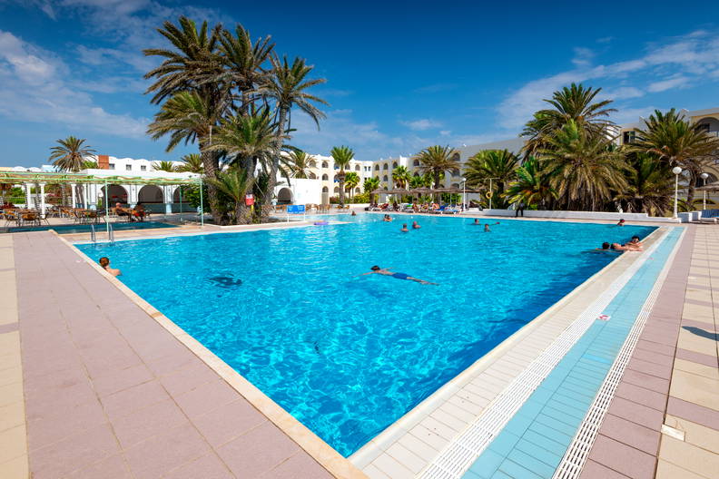 Basen w hotelu otoczony palmami i wypoczywającymi osobami.