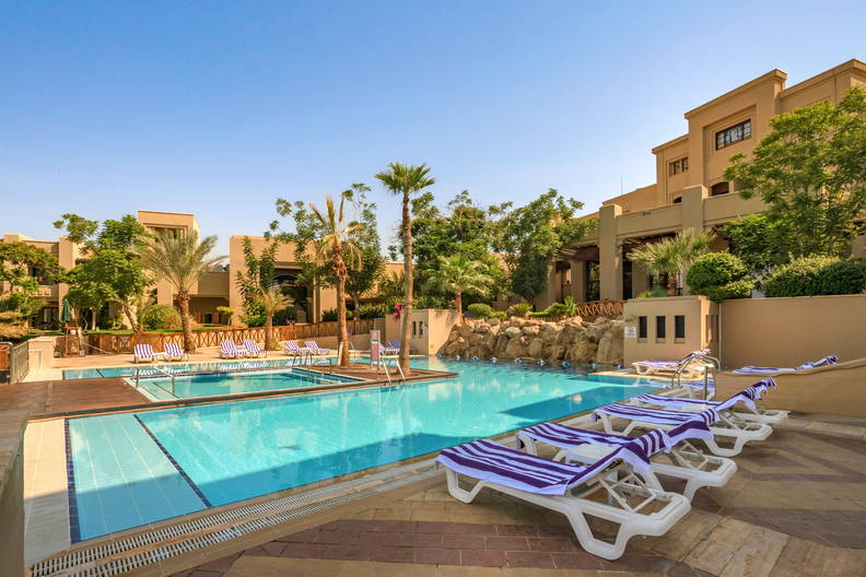 Basen z leżakami na tle hotelu i zieleni.