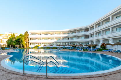 Basen hotelowy z leżakami i białym budynkiem w tle.