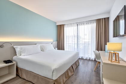 Jasny pokój hotelowy z dużym łóżkiem, biurkiem i balkonem.