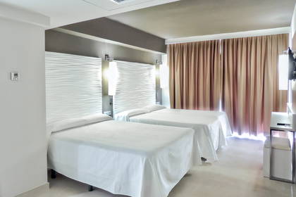 Pokój hotelowy z dwoma łóżkami, stolikiem i zasłonami w oknach.