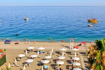 Plaża z białymi parasolami i leżakami. W tle morze z łodziami.